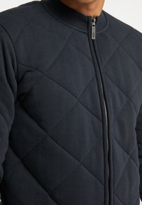 Quiltad mörkblå jacka med hög krage, dragkedjestängning och diamantmönster. Tillverkad av mjukt material med en slät yta.