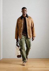 Brun, knap-op jakke med en sort krave, brun skjorte under, olivengrønne cargo-bukser, beige sko og en hvid håndtaske med brune detaljer.