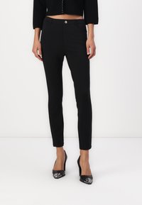 Pantalones ajustados negros con una textura suave, silueta ajustada, bolsillos frontales y cierre de botón dorado. Combinados con tacones negros de punta.