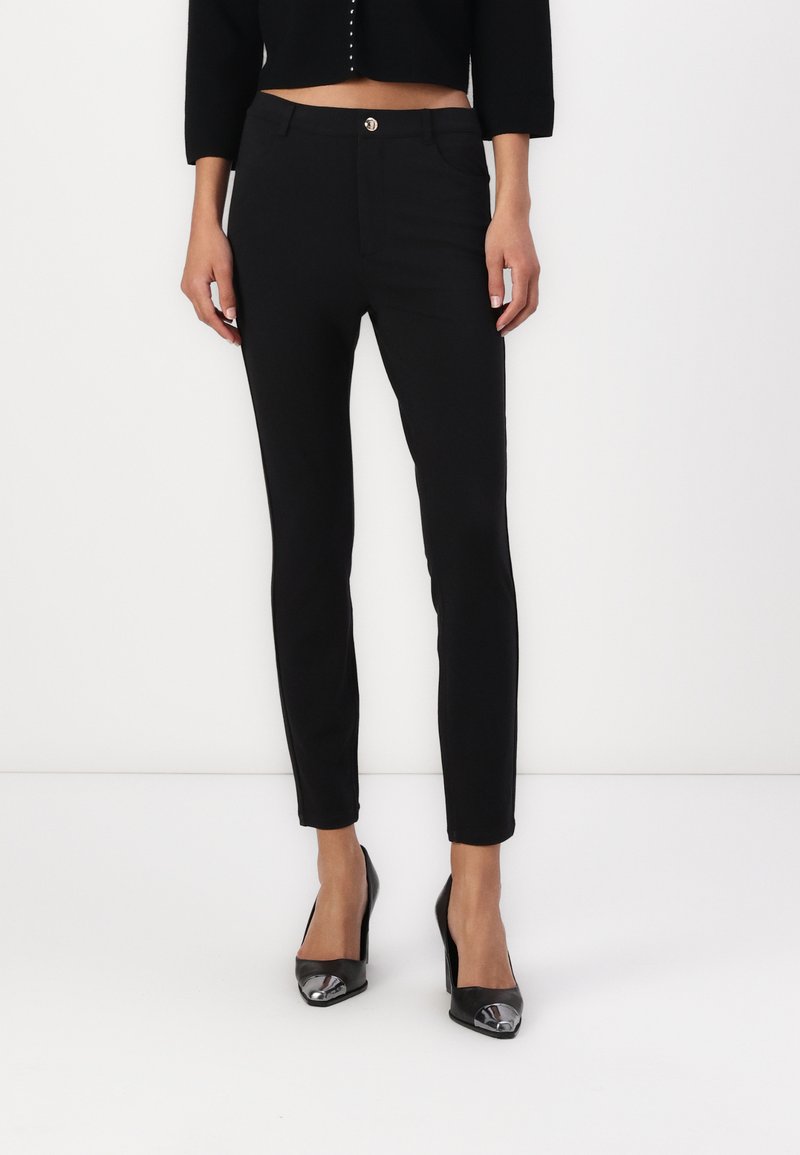 Pantalones ajustados negros con una textura suave, silueta ajustada, bolsillos frontales y cierre de botón dorado. Combinados con tacones negros de punta.