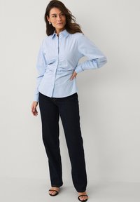 Chemise boutonnée bleu clair avec détails froncés à la taille, associée à un jean en denim foncé et des sandales noires à talons.