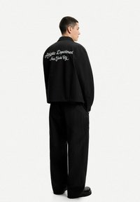Black cropped jacket, features embroidered text on the back; paired with loose black pants and chunky black boots. Matte texture, oversized fit.