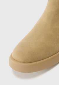 Botín de ante en beige claro, punta redondeada, textura suave y suela de goma. Presenta un diseño sencillo con mínimos acentos y costuras.