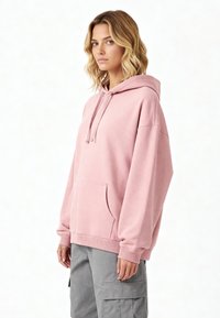 Felpa rosa oversize realizzata in tessuto morbido, con tasca a marsupio, cappuccio con coulisse e polsini a coste. Abbinata a pantaloni cargo grigi.