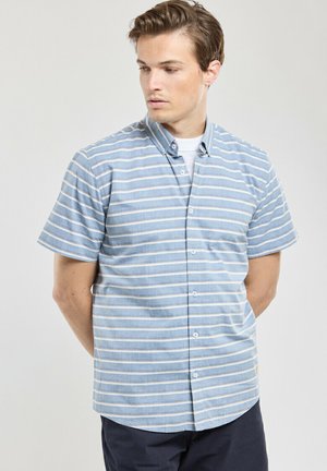 Homme aux cheveux châtain clair portant une chemise boutonnée à manches courtes rayée bleu et blanc, debout les mains derrière le dos.