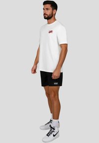 Camiseta de algodón blanca con mangas cortas, pequeño logotipo rojo en el pecho, combinada con pantalones cortos negros y zapatillas blancas con acentos negros.