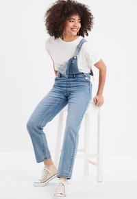 Pantalones de mezclilla con tirantes ajustables, color azul claro y piernas ajustadas. Combinados con una camiseta blanca y zapatillas beige.