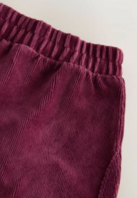Burgundské kordové joggingové kalhoty s vroubkovaným elastickým pasem a texturovanou tkaninou, která má vertikální žebrování a jemné povrchové detaily.
