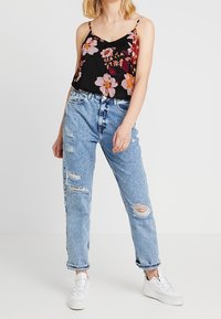 Camisole noire à motifs floraux avec bretelles spaghetti, associée à un jean bleu clair déchiré. Le mannequin porte des baskets blanches. Détails lumineux à motif floral.