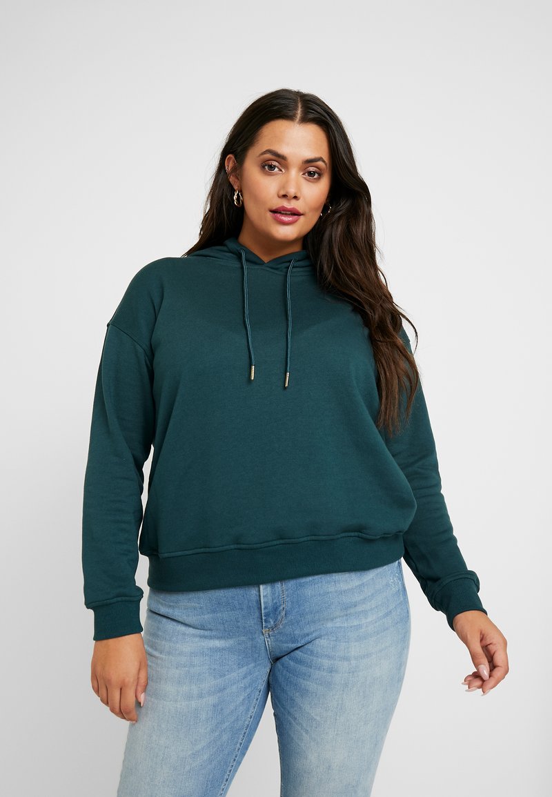 Teal hettegenser med løs passform, snøring, ribbede mansjetter og kant. Kombinert med lyseblå jeans for en avslappet look.