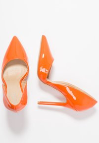 Chaussures à talons hauts en cuir verni orange avec bouts pointus, présentant un design découpé et un talon aiguille fin. Texture lisse et brillante.