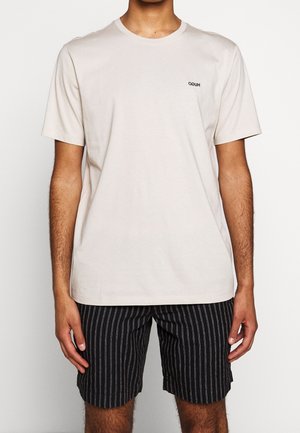 T-shirt en coton beige clair avec un col rond et des manches courtes, arborant un petit logo noir sur la poitrine. Portée avec un short à rayures noires.