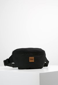 Marsupio in tessuto nero con chiusura a zip, tracolla regolabile e una patch rettangolare in pelle marrone. Texture liscia, design compatto.