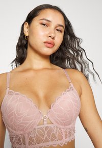 Bralette de renda em rosa suave com padrão floral, decote em V, alças ajustáveis e suporte com aros. Apresenta um detalhe em forma de chave ao centro.