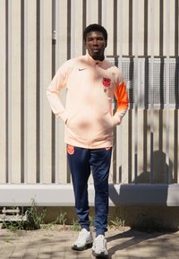 Light pink Nike jacket with orange accents and FC Barcelona logo, paired with navy pants. White sneakers and a textured backdrop.