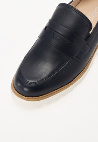 Mocassin en cuir noir avec surpiqûres marron sur la bordure et semelle blanche, doté d'un design à enfiler et d'une bride décorative à l'avant.