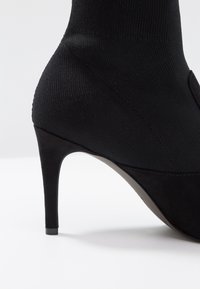 Botín negro de tacón alto con parte superior de punto similar a un calcetín y un acabado de ante suave en la punta. Presenta un tacón delgado y puntiagudo.