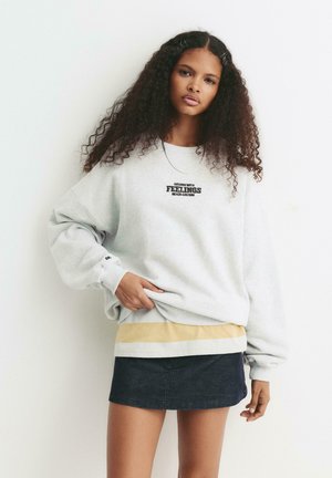 Sweatshirt gris avec le texte "GRANDI AVEC DES ÉMOTIONS", manches à patte, et un bas superposé rayé jaune par-dessus une jupe en denim foncé.