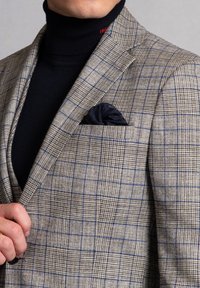 Blazer a quadri in tonalità beige, marrone e blu. Presenta un fazzoletto da taschino in blu navy, un dolcevita nero sotto, tessuto testurizzato e rever classici.