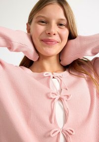 Różowy sweter z prążkowanej dzianiny z otwartym przodem, ozdobiony dwoma kokardkami i długimi rękawami. Miękka tekstura, nieco luźny krój, nałożony na biały top.