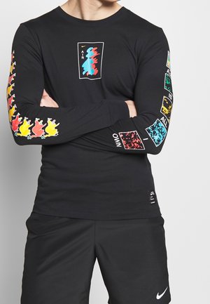 Personne portant un t-shirt noir à manches longues avec des motifs abstraits colorés sur la poitrine et les manches, assorti à un short noir Nike.