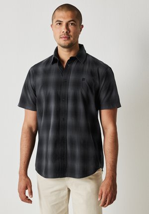 Chemise à manches courtes en motif tartan noir et gris, fabriquée dans un tissu lisse, avec un col boutonné et un petit logo sur la poitrine.