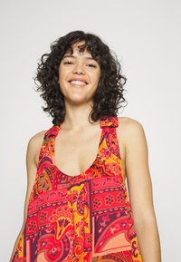 Vestido sem mangas colorido com um decote profundo. Apresenta um padrão paisley em tons de vermelho, laranja e amarelo. O tecido parece leve e fluido.
