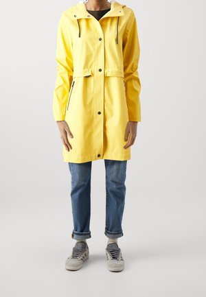 Vista frontal de una persona que lleva un impermeable con capucha de color amarillo brillante, vaqueros azules remangados y zapatillas grises sobre un fondo liso.