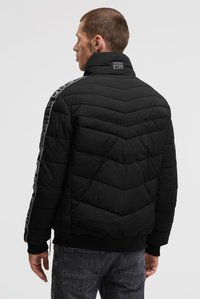 Mann trägt eine schwarze gesteppte Jacke mit Logo-Schriftzug am Kragen und Ärmel, steht mit dem Rücken zu uns vor einem einfarbigen hellen Hintergrund.