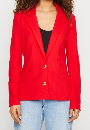 Frau trägt einen leuchtend roten Blazer mit goldenen Knöpfen über einem schwarzen Top und hellblauen Jeans, steht vor einem schlichten Hintergrund.