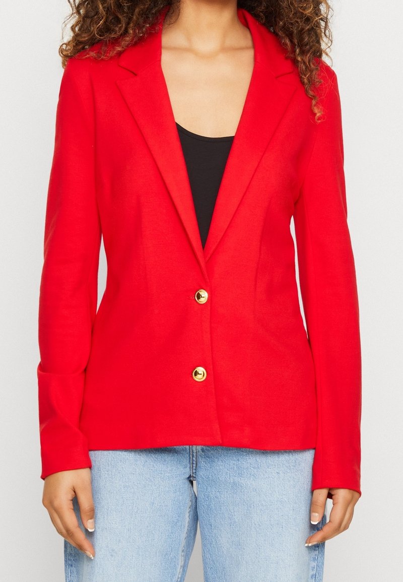 Femme portant un blazer rouge vif avec des boutons dorés sur un haut noir et un jean bleu clair, debout devant un fond uni.