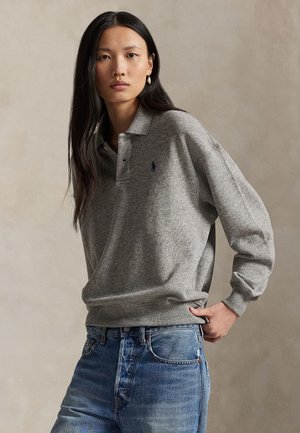 Kobieta z długimi czarnymi włosami, ubrana w szary sweter z kołnierzem i niebieskie dżinsy, stoi z jedną ręką w kieszeni na jednolitym tle.