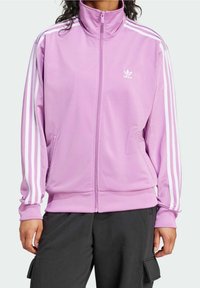 Person som bär en lavendelfärgad Adidas zip-up träningsjacka med vita ränder på ärmarna och svarta byxor med sidofickor.