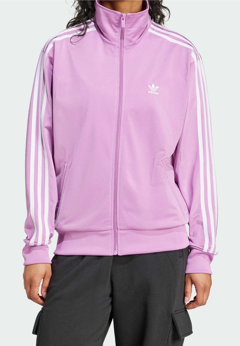 Person som bär en lavendelfärgad Adidas zip-up träningsjacka med vita ränder på ärmarna och svarta byxor med sidofickor.