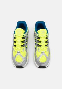 Heldere gele sportschoenen van mesh en synthetisch materiaal, met zilveren accenten, zwarte veters en een blauwe binnenvoering. Klassiek ontwerp met drie strepen.