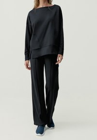 Czarna bluza z długim rękawem, szerokim dekoltem i opuszczonymi ramionami, zestawiona z luźnymi czarnymi spodniami i niebieskimi butami sportowymi.