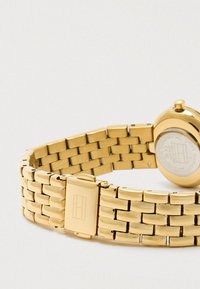 Złota bransoletka na zegarek z ogniwami i zapięciem z prostokątnym logo, przymocowana do okrągłej koperty zegarka z wygrawerowanym napisem.