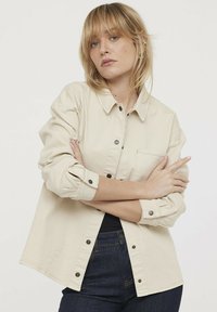 Camisa beige de botones con un bolsillo, mangas largas arremangadas y una tela texturizada. Botones negros acentúan el cierre frontal.