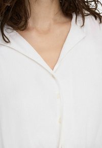 Chemisier blanc à boutons, en tissu doux et texturé, avec un col pointu et trois boutons sur le devant.