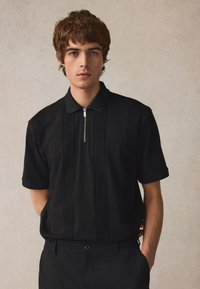 Černé polo tričko se zipovým límečkem, zdobené texturovanými vertikálními vzory. V kombinaci s černými kalhotami, což vytváří monochromatický vzhled.