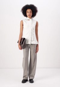 Weiße ärmellose Bluse mit Bindegürtel, kombiniert mit grauen weit geschnittenen Hosen und schwarzen Slippern. Hält eine braune Clutch.