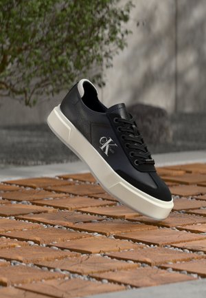 Zapatilla negra de cuero con suela blanca y logotipo "cK", flotando sobre un suelo de baldosas de madera al aire libre junto a un árbol y una pared de hormigón.
