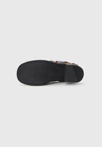 Suela de goma negra con agarre texturizado, unida a una parte superior de cuero marrón con una correa ajustable de hebilla. Diseño minimalista.