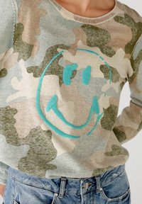Pullover in maglia con un motivo mimetico in tonalità beige, verde e blu; presenta un grande sorriso ricamato turchese al centro.