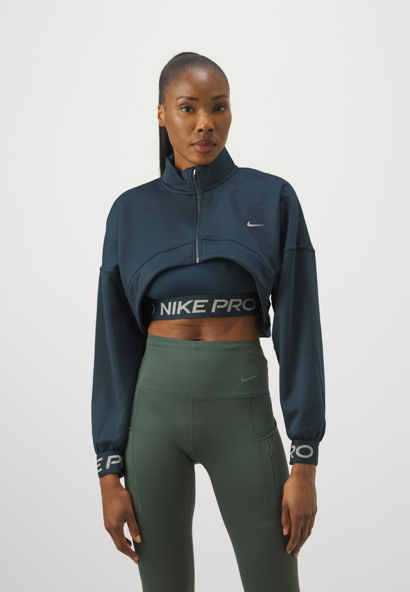 Mornarska skraćena jakna na zatvarač s visokim ovratnikom, sadrži Nike logo; uparena s tamnozelenim tajicama visokog struka i odgovarajućim sportskim grudnjakom.