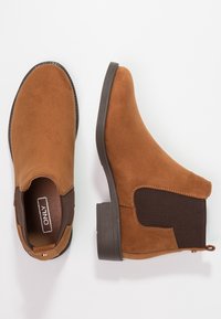 Botas Chelsea de ante marrón con paneles laterales elásticos y tacones bajos apilados, mostradas en vistas superior y lateral sobre un fondo blanco.