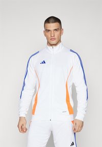 Hvid atletisk jakke med blå og orange detaljer, lynlås fortil, ribbede manchetter og tre-stribet design på skuldrene.