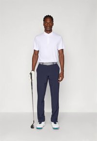 Homem em pé segurando um taco de golfe com a mão direita, vestindo uma camisa polo branca, calças azul-marinho, sapatos de golfe brancos e uma luva de golfe branca na mão esquerda.