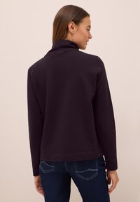 Haut long à manches violettes foncées avec un col montant, texture lisse et coupe décontractée, associé à un jean en denim foncé avec des poches arrière.