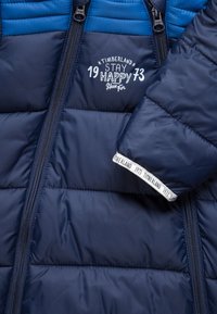 Marinblå vadderad jacka med horisontell sömnad, en blå quiltad ok, dubbla dragkedjor och vit tryckt logotyp nära fållen.